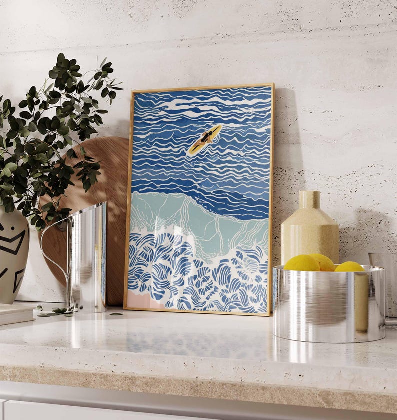 May include: A framed art print depicts a person on a yellow surfboard in blue ocean waves. The artwork has a gold frame and features a beach scene with white, blue, and light blue colors. The print is displayed on a white countertop.