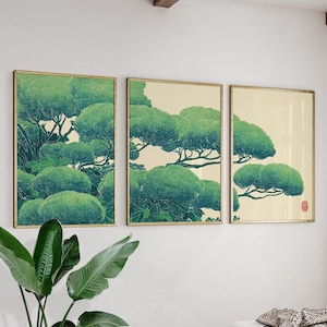 May include: Three framed art prints showcasing a stylized depiction of green trees against a cream backdrop. The artwork, with a Japanese aesthetic, features a minimalist style and gold frames. The prints are displayed on a white wall.