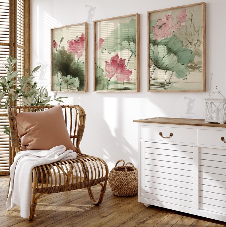May include: Three framed art prints featuring watercolor paintings of pink lotus flowers and green leaves. The artwork has a vintage aesthetic, with a light beige background and wooden frames. The prints are displayed on a white wall, above a white cabinet.