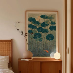 May include: A framed art print featuring a teal and green lotus pond scene with a small orange fish. The artwork is displayed above a wooden cabinet with a round beige vase and a small lamp.