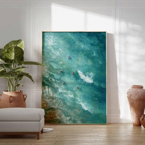 May include: Large framed art print depicting an aerial view of ocean waves and several people swimming. The artwork is in shades of blue and green, with a gold frame. The print is displayed in a room with a plant, ottoman, and decorative vases.