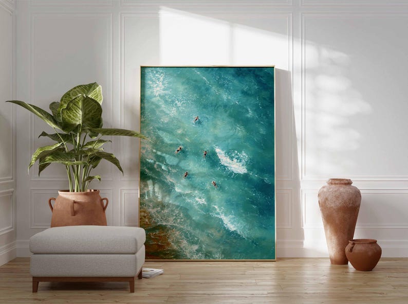 May include: Large framed art print depicting an aerial view of ocean waves and several people swimming. The artwork is in shades of blue and green, with a gold frame. The print is displayed in a room with a plant, ottoman, and decorative vases.