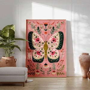 May include: A framed art print featuring a large butterfly with intricate floral patterns in pink, green, and cream colors. The artwork is set against a pink background, with a wooden frame. The print is displayed in a room with a plant, ottoman, and decorative vases.
