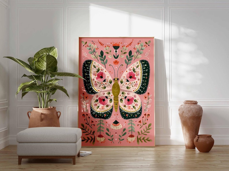 May include: A framed art print featuring a large butterfly with intricate floral patterns in pink, green, and cream colors. The artwork is set against a pink background, with a wooden frame. The print is displayed in a room with a plant, ottoman, and decorative vases.