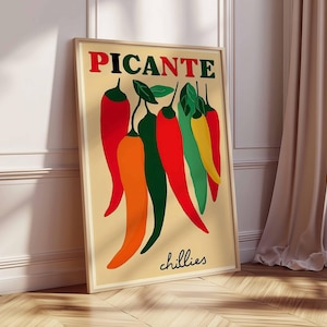 May include: A framed poster with the word "PICANTE" in red and green above a collection of stylized chili peppers in red, orange, green, and yellow. The word "chillies" is written in cursive at the bottom of the poster. The poster is leaning against a white wall.