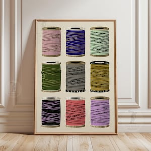 Puede incluir: Una impresión enmarcada con nueve carretes de hilo en varios colores, incluyendo rosa, azul, verde, gris y negro. Cada carrete tiene una base de madera y está envuelto con hilo de un color contrastante. La obra de arte tiene un fondo neutro y un marco de madera.