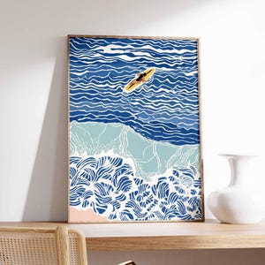 May include: A framed art print depicting a person on a yellow surfboard in blue ocean waves. The artwork features shades of blue and white, with a pink section representing the beach. The print is displayed on a wooden shelf.
