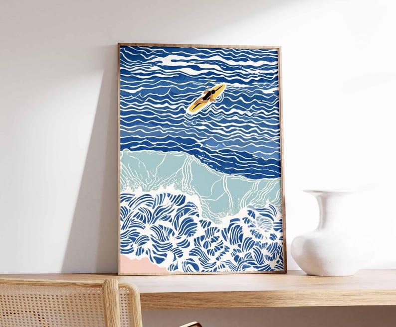 May include: A framed art print depicting a person on a yellow surfboard in blue ocean waves. The artwork features shades of blue and white, with a pink section representing the beach. The print is displayed on a wooden shelf.