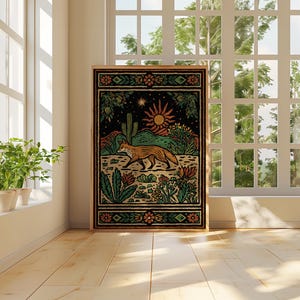 May include: A framed art print featuring a desert landscape with a fox, cactus, and sun. The artwork has a black background with green, orange, and yellow details. The print is displayed in a room with a window and plants.