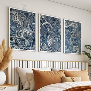 May include: Three framed abstract art prints with a navy blue and white swirling design hang above a bed. The artwork is displayed above a bed with orange and cream-colored pillows and a wooden headboard.