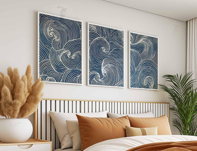 May include: Three framed abstract art prints with a navy blue and white swirling design hang above a bed. The artwork is displayed above a bed with orange and cream-colored pillows and a wooden headboard.