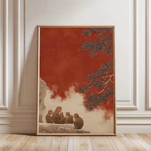 May include: A framed art print depicting monkeys in a traditional Japanese style. The image shows monkeys with red faces, set against a warm red backdrop with a pine tree branch and white clouds. The frame is a light brown colour.