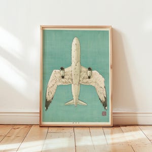 May include: A framed art print featuring a top-down view of a white aeroplane with wings that transition into feathered bird wings. The aircraft is centred against a solid teal background, with small red text and a red stamp in the lower corner.