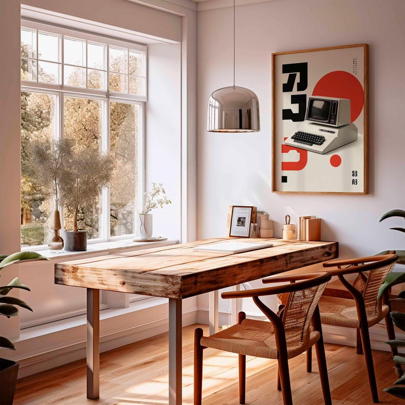May include: A bright, airy room with a wooden desk and two chairs. A framed poster with a vintage computer graphic hangs on the wall. A large window offers a view of trees. A silver pendant light hangs above the desk.