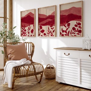 May include: Three framed wall art pieces with a red and beige watercolor landscape design. The artwork features mountain silhouettes and floral elements. The frames are a light wood color, and the overall aesthetic is modern and minimalist.