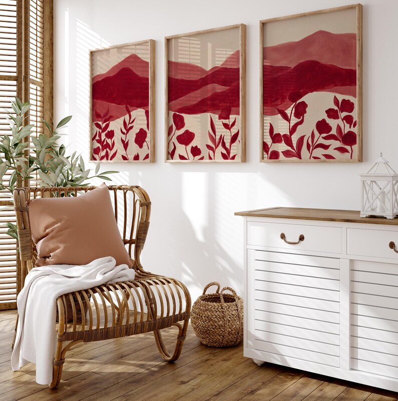 May include: Three framed wall art pieces with a red and beige watercolor landscape design. The artwork features mountain silhouettes and floral elements. The frames are a light wood color, and the overall aesthetic is modern and minimalist.