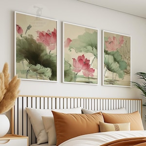 May include: Three framed art prints featuring watercolor paintings of pink lotus flowers and green leaves. The artwork is displayed above a bed with orange and white pillows. The prints are in white frames.