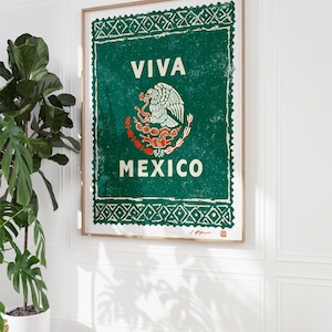May include: Framed art print featuring the Mexican coat of arms and the words "VIVA MEXICO" in white on a green background. The print has a distressed, vintage look and is displayed on a white wall next to a potted plant.