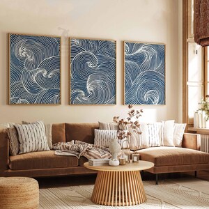 May include: Three framed wall art pieces with a blue and white wave design. The artwork is displayed above a brown sofa with decorative pillows. A round wooden coffee table and a woven rug complete the living room setting.