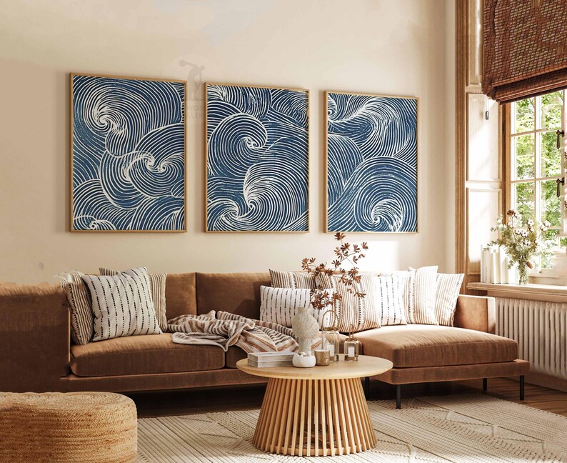 May include: Three framed wall art pieces with a blue and white wave design. The artwork is displayed above a brown sofa with decorative pillows. A round wooden coffee table and a woven rug complete the living room setting.