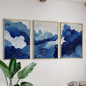 May include: Set of three framed wall art pieces. Each piece features an abstract design of swirling blue and white lines, resembling clouds or waves. The frames are gold-colored, and the artwork is displayed against a white wall.
