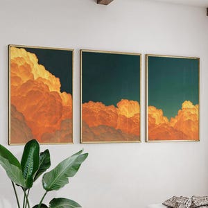 May include: Three framed art prints depicting a cloudscape with orange and yellow clouds against a dark teal sky. The artwork is in gold frames and mounted on a white wall. A green plant is visible in the lower left corner.
