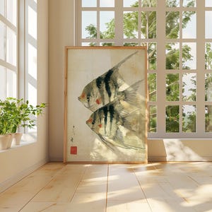 K&ouml;nnte beinhalten: Ein gerahmter Aquarelldruck mit zwei stilisierten Fischen mit schwarz-wei&szlig;en Streifen und roten Akzenten. Das Kunstwerk wird in einem hellen Raum mit einem Fenster und Pflanzen ausgestellt, was eine ruhige Atmosph&auml;re schafft. Der Druck hat einen Holzrahmen.