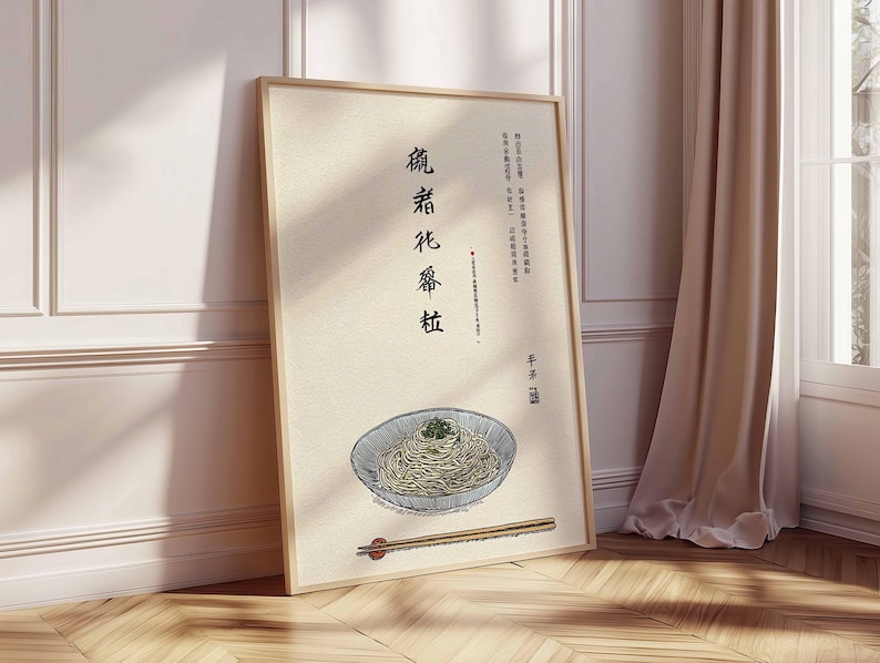 May include: A framed art print featuring a bowl of noodles with chopsticks. The artwork has a beige background with Chinese characters and a bowl of noodles with green garnish. The frame is light brown, and the print is leaning against a white wall and wooden floor.