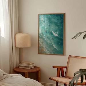 May include: A framed art print depicts an aerial view of ocean waves in shades of turquoise and white. Several small figures are visible in the water, suggesting surfers or swimmers. The artwork is displayed on a neutral-colored wall, adding a touch of coastal decor.