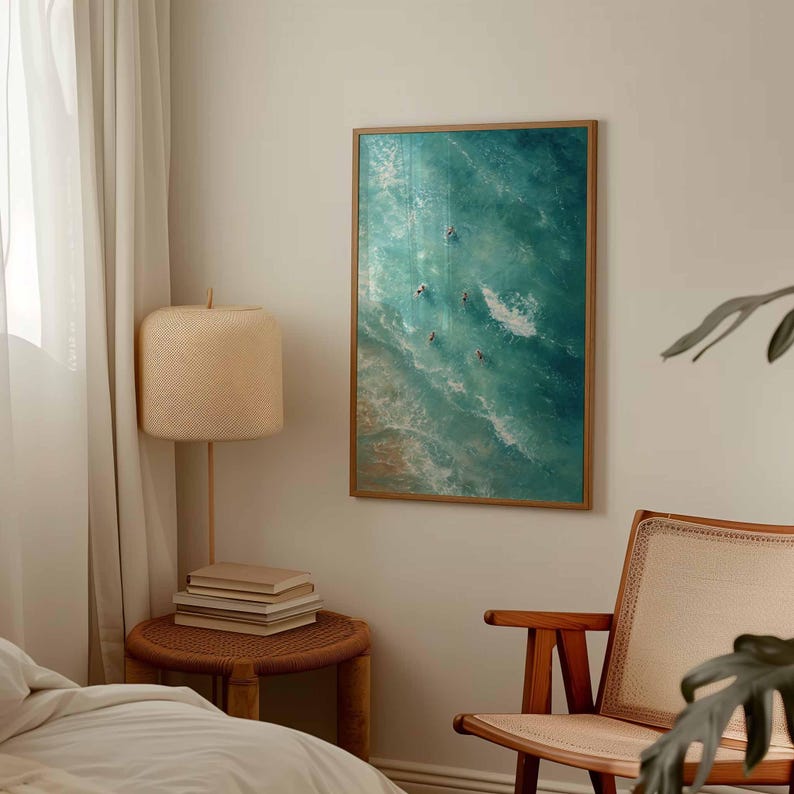 May include: A framed art print depicts an aerial view of ocean waves in shades of turquoise and white. Several small figures are visible in the water, suggesting surfers or swimmers. The artwork is displayed on a neutral-colored wall, adding a touch of coastal decor.