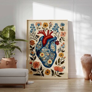May include: A framed art print featuring a stylized anatomical heart design. The heart is blue with floral patterns and red arteries, surrounded by colorful flowers. The artwork is displayed in a room with a plant, ottoman, and decorative vases.