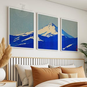 K&ouml;nnte beinhalten: Drei gerahmte Kunstdrucke, die eine Bergkette in Blau- und Wei&szlig;t&ouml;nen zeigen. Das Kunstwerk hat einen minimalistischen Stil mit hellblauem Himmel und wei&szlig;en R&auml;ndern. Die Drucke werden &uuml;ber einem Bett mit orangefarbenen Kissen angezeigt.
