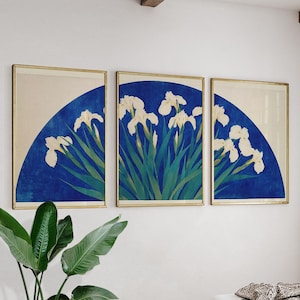May include: Set of three framed art prints featuring white iris flowers against a blue background. The artwork has a gold frame and a cream-colored upper section. The iris flowers have green leaves.