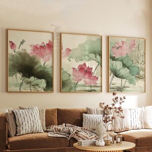 May include: Three framed watercolor paintings of pink lotus flowers and green lily pads. The artwork is in a light-colored frame. The paintings are displayed above a brown sofa with striped pillows and a round wooden coffee table.