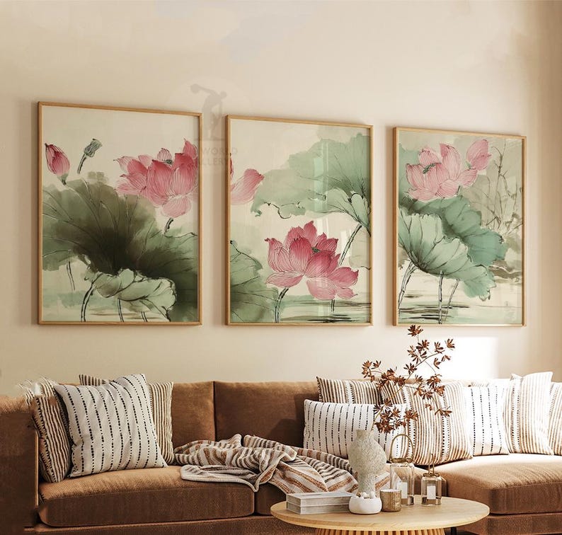 May include: Three framed watercolor paintings of pink lotus flowers and green lily pads. The artwork is in a light-colored frame. The paintings are displayed above a brown sofa with striped pillows and a round wooden coffee table.
