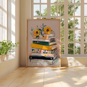 May include: A framed art print featuring a stack of books with a floral arrangement of sunflowers and other flowers on top. The books are various colors, including yellow, green, and floral patterns. The artwork is displayed in a room with a window and plants.
