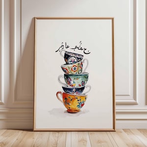 Puede incluir: Impresión en acuarela enmarcada de una pila de cinco tazas de té con diseños florales en varios colores, incluyendo azul, naranja y verde. Las tazas están apiladas una encima de la otra. La impresión está en un marco de madera marrón claro. Texto árabe en la parte superior.
