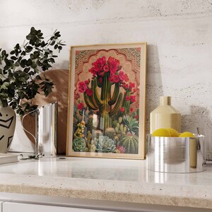 May include: A vintage-style print of a blooming cactus with red flowers in a gold frame. The print is on a white wall with a wooden cutting board and a silver pitcher in the foreground.