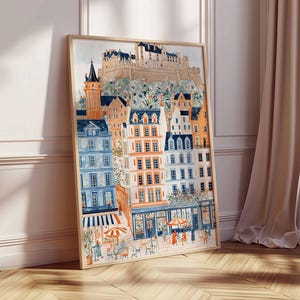 May include: A framed watercolour print of a European city scene. The artwork depicts colourful buildings with orange, blue, and white facades, a castle on a hill, and people at outdoor cafes. The print is displayed on a wooden floor.
