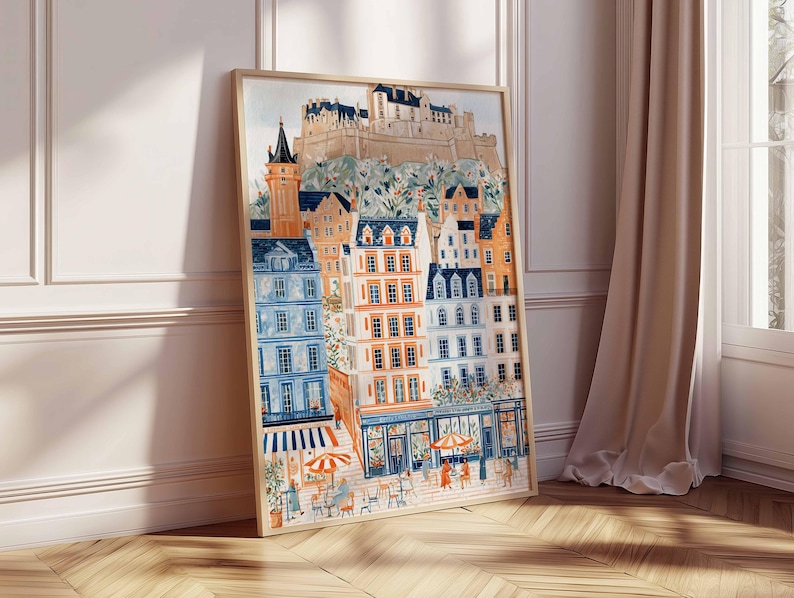 May include: A framed watercolour print of a European city scene. The artwork depicts colourful buildings with orange, blue, and white facades, a castle on a hill, and people at outdoor cafes. The print is displayed on a wooden floor.