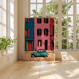 May include: A framed art print depicts a colorful street scene with a teal car. The buildings are painted in vibrant hues of pink and teal, with black window shutters and orange doors. The artwork is displayed in a bright room with large windows and potted plants.