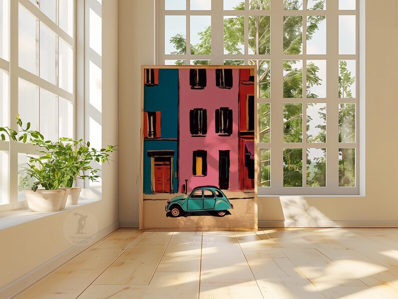 May include: A framed art print depicts a colorful street scene with a teal car. The buildings are painted in vibrant hues of pink and teal, with black window shutters and orange doors. The artwork is displayed in a bright room with large windows and potted plants.