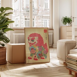 May include: A framed print featuring a colorful, stylized lion-dog illustration. The artwork showcases a red and pink mane, floral patterns, and traditional Japanese text. The print is displayed on a wooden floor, leaning against a wall.