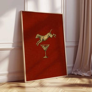 Pu&ograve; includere: Stampa artistica incorniciata con un cane dorato che salta sopra un bicchiere da martini dorato su uno sfondo rosso strutturato. L'opera d'arte &egrave; in una cornice dorata e poggia su un muro bianco e un pavimento in legno.