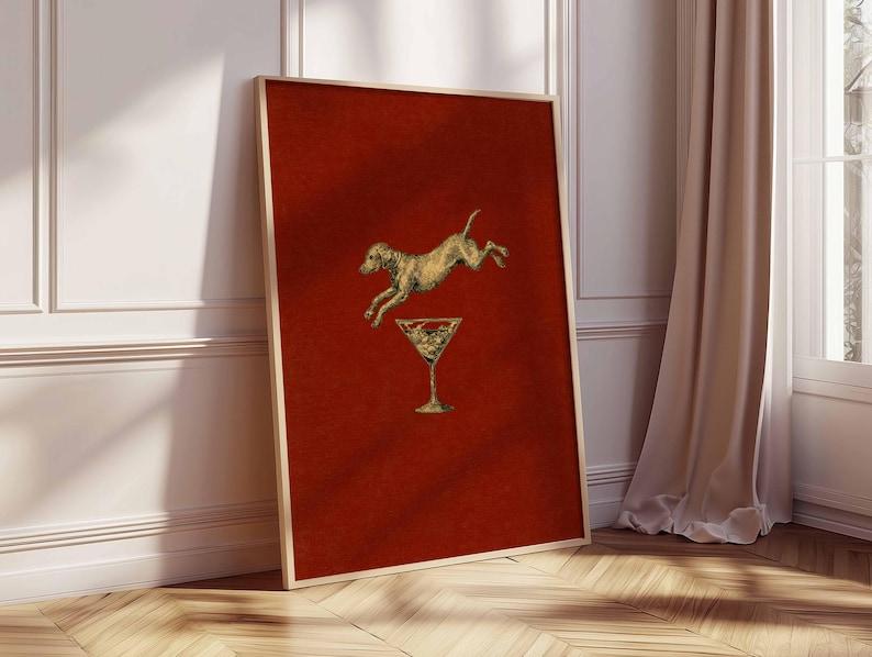 Pu&ograve; includere: Stampa artistica incorniciata con un cane dorato che salta sopra un bicchiere da martini dorato su uno sfondo rosso strutturato. L'opera d'arte &egrave; in una cornice dorata e poggia su un muro bianco e un pavimento in legno.