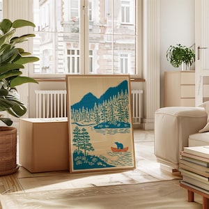 May include: A framed art print featuring a teal and orange illustration of a bear in a canoe on a lake, with mountains and trees in the background. The print is in a wooden frame and leans against a cardboard box.