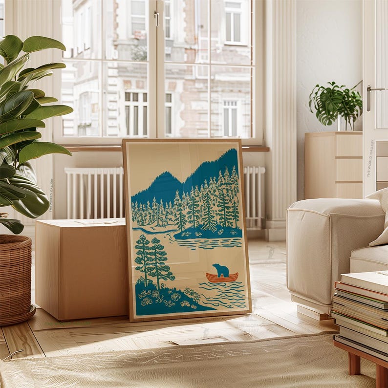 May include: A framed art print featuring a teal and orange illustration of a bear in a canoe on a lake, with mountains and trees in the background. The print is in a wooden frame and leans against a cardboard box.