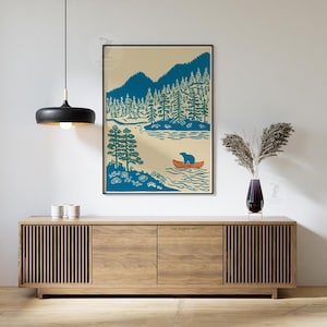 May include: A framed art print featuring a blue bear in an orange canoe on a lake, surrounded by blue mountains and green trees. The artwork has a vintage, outdoorsy theme, perfect for a cabin or nature-themed decor.