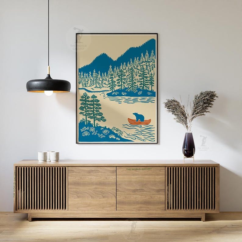 May include: A framed art print featuring a blue bear in an orange canoe on a lake, surrounded by blue mountains and green trees. The artwork has a vintage, outdoorsy theme, perfect for a cabin or nature-themed decor.