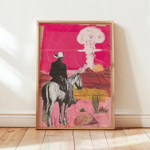 May include: Framed art print featuring a cowboy on horseback in a desert landscape with a mushroom cloud in the background. The artwork has a pink and red color scheme with a wooden frame. The cowboy is wearing a white hat and black coat.